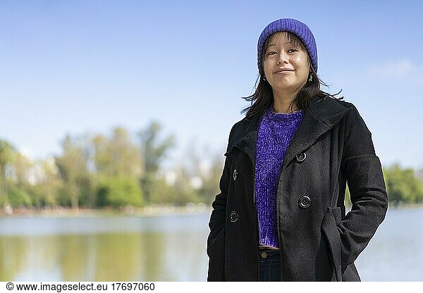 Portrait of a young Latin woman dressed in a pullover  coat and wool cap  looking at the camera  at a lake with copy space
