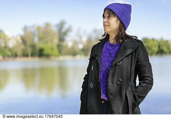 Portrait of a young Latin woman dressed in a pullover  coat and wool cap  looking to the side  at a lake with copy space