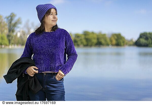 Portrait of a young Latin woman dressed in a pullover  coat and wool cap  looking to the side  at a lake with copy space