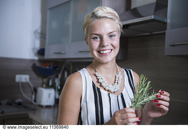 Portrait of a young woman holding rosemary in the kitchen and smiling