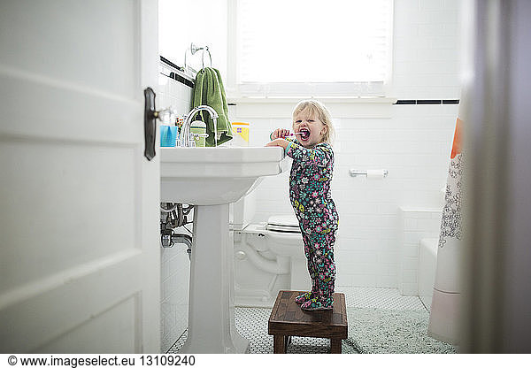Portrait of baby girl brushing teeth while standing on stool in bathroom