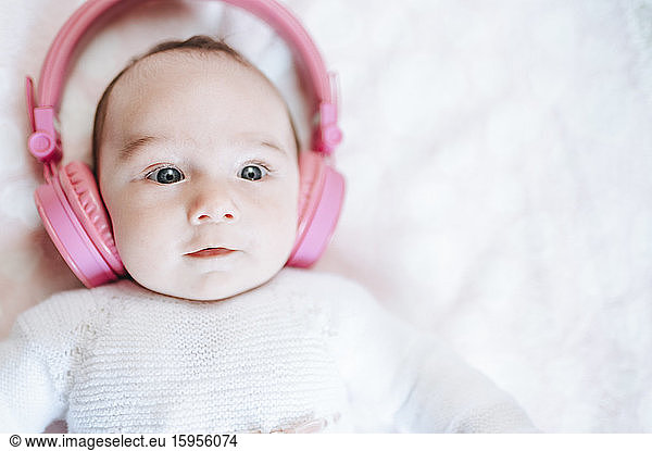 Portrait of baby girl with oversized pink headphones lying on white blanket