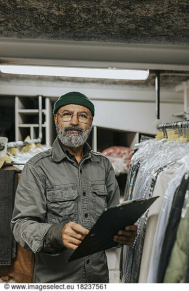 Portrait of bearded tailor with clipboard standing by clothing rack in workshop