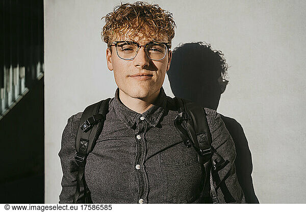 Portrait of blond male student wearing eyeglasses holding book against gray wall