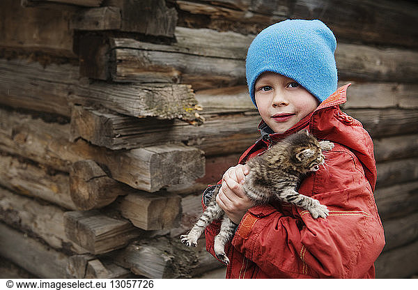 Portrait of boy carrying cat while standing against wooden wall