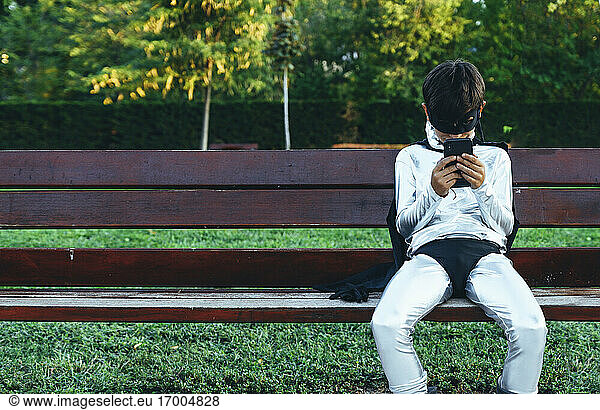 Portrait of boy disguised as superhero using mobile phone on park bench