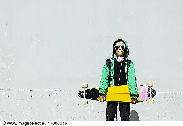 Portrait of boy with skateboard and headphones standing in front of white wall