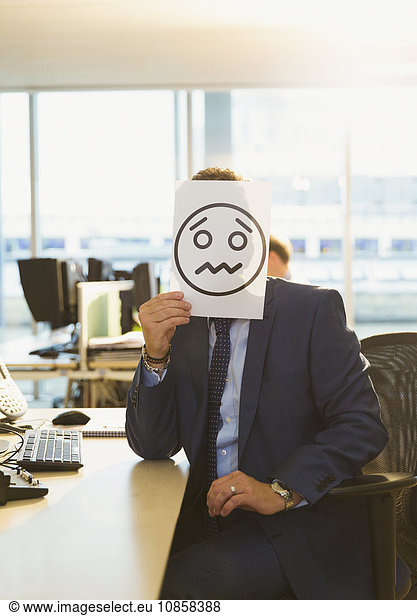 Portrait of businessman holding frowning face printout over his face in office