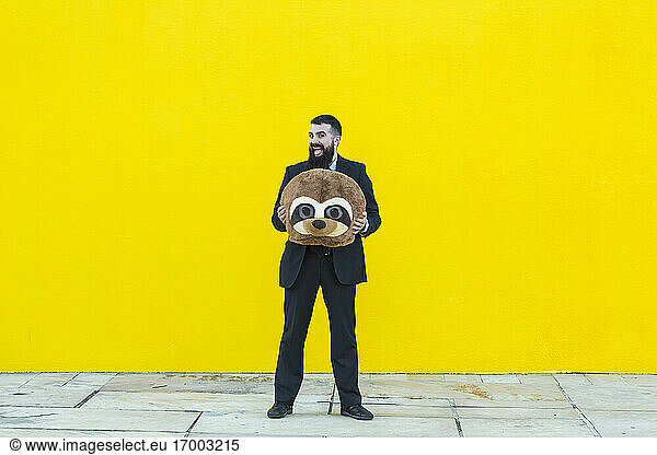 Portrait of businessman in black suit holding meerkat mask in front of yellow wall