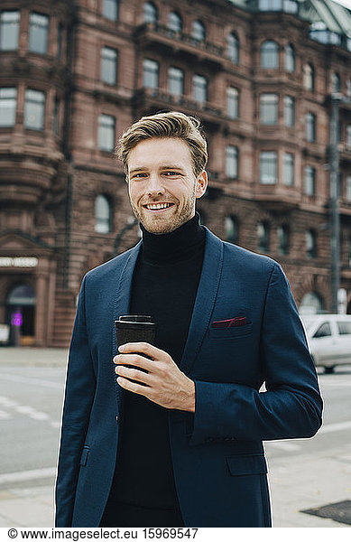 Portrait of businessman with disposable cup standing against building in city