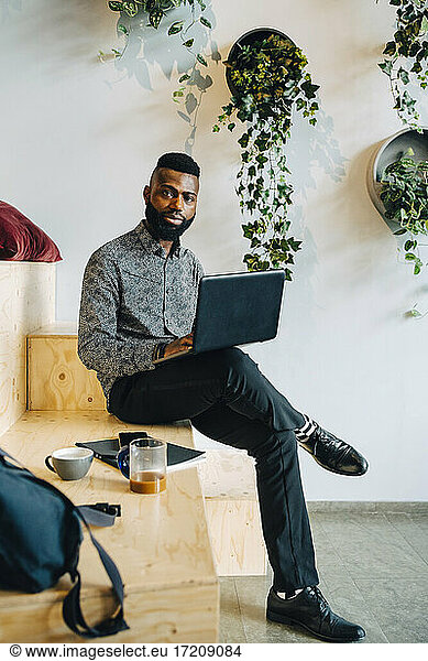 Portrait of businessman with laptop sitting on wooden seat in office