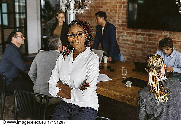 Portrait of businesswoman standing with arms crossed while colleagues discussing in background