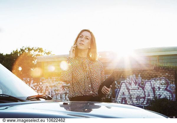 Portrait of businesswoman with file standing behind parked car telephoning at backlight
