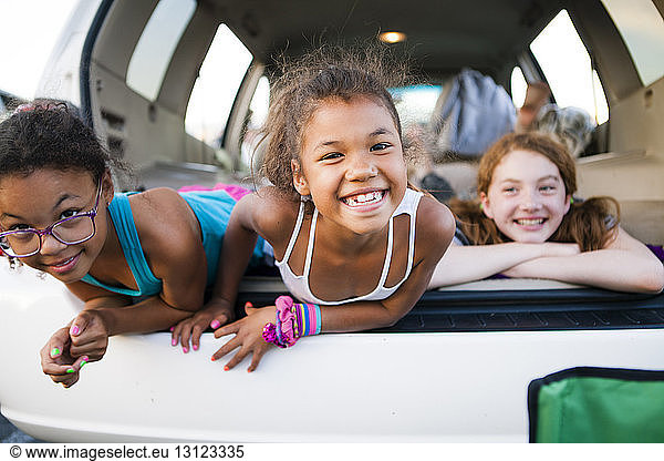 Portrait of carefree sisters lying in car trunk with friend