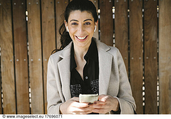 Portrait of cheerful businesswoman holding smart phone against wooden wall