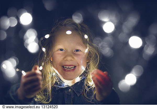Portrait of cheerful girl playing with illuminated string lights during night