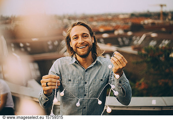 Portrait of cheerful man holding string light while standing on terrace in city