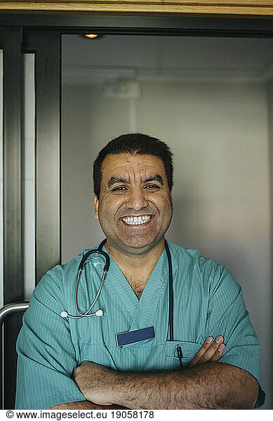 Portrait of cheerful mature doctor standing with arms crossed in front of door at hospital