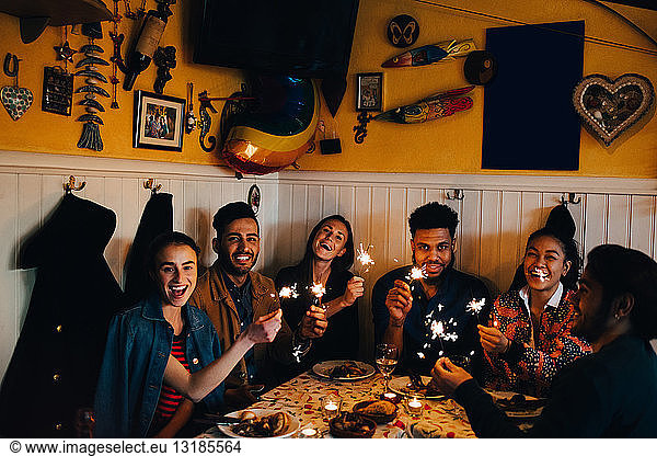Portrait of cheerful young multi-ethnic friends sitting with burning sparklers at table in restaurant