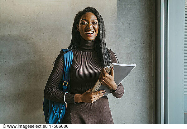 Portrait of cheerful young woman holding book standing with backpack against gray wall