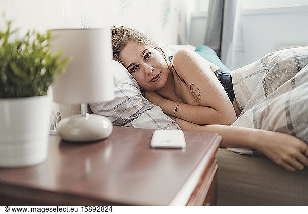 Portrait of confident attractive young woman lying in bed at home