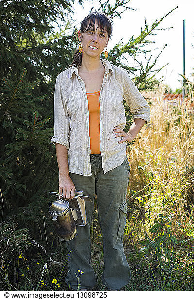 Portrait of confident beekeeper with hand on hip holding bee smoker while standing against plants at field