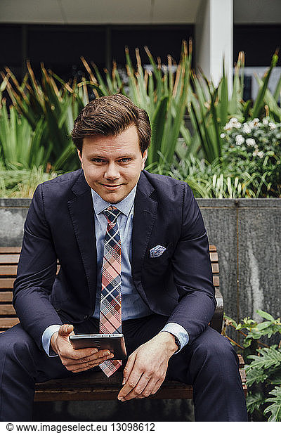 Portrait of confident businessman holding tablet computer while sitting on sidewalk bench