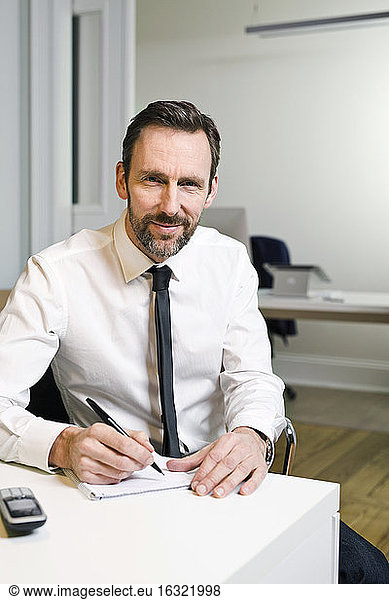 Portrait of confident businessman sitting at desk in office taking notes