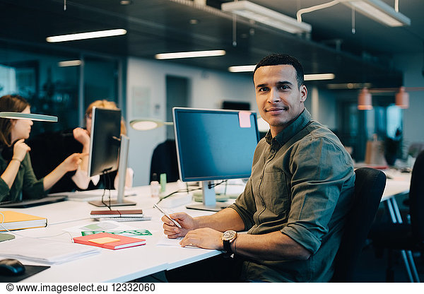 Portrait of confident businessman sitting at desk while female colleagues working in creative office