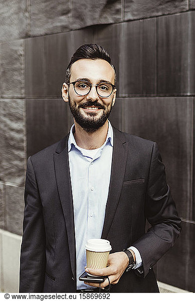 Portrait of confident businessman with disposable cup standing against wall