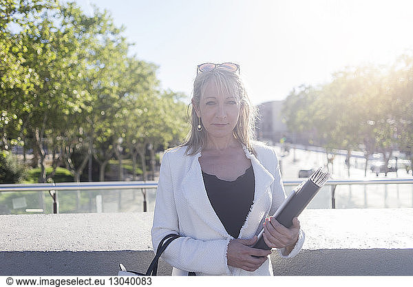 Portrait of confident businesswoman holding file while standing in city during sunny day