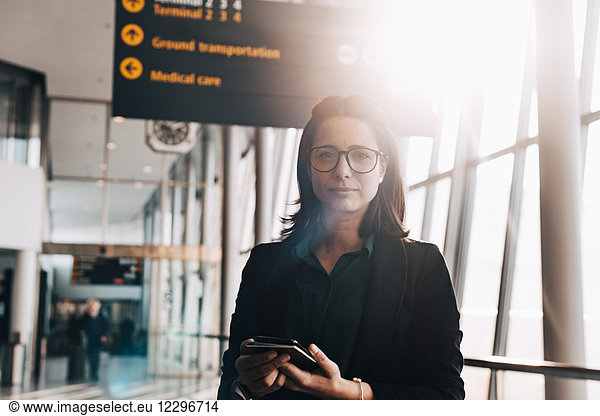 Portrait of confident businesswoman holding mobile phone and passport in airport terminal