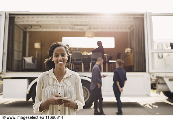 Portrait of confident businesswoman holding smart phone with colleagues and portable office truck on road in background