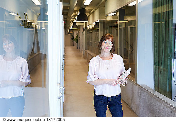 Portrait of confident businesswoman holding spiral notebook while standing at corridor in office