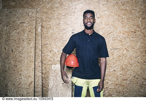 Portrait of confident carpentry student holding hardhat while standing against wooden wall