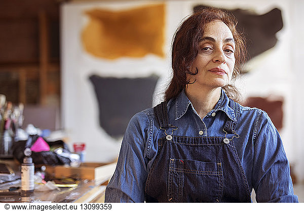 Portrait of confident female artist standing against paintings in workshop