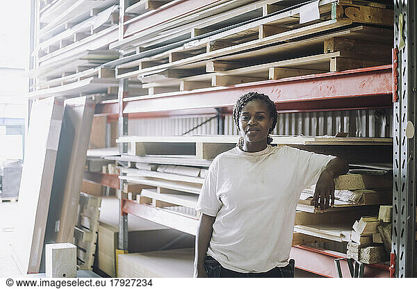 Portrait of confident female carpenter standing by pallet stack in warehouse