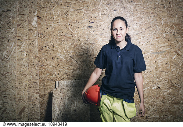 Portrait of confident female carpentry student holding hardhat while standing against wooden wall
