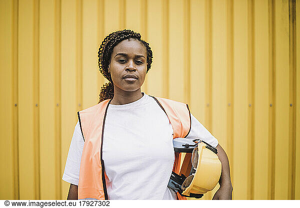 Portrait of confident female construction worker with hardhat against yellow metal wall