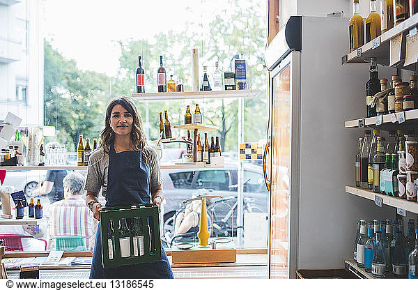 Portrait of confident female employee carrying bottles in crate at deli