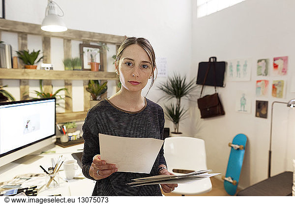 Portrait of confident female illustrator holding papers while standing in creative office