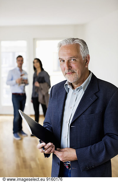 Portrait of confident male real estate agent with couple standing in background at home