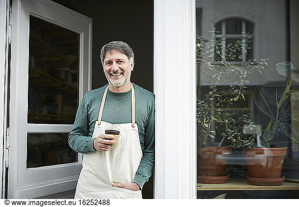 Portrait of confident mature male owner having coffee at doorway of art studio