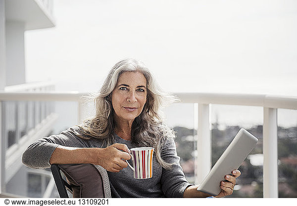 Portrait of confident mature woman holding tablet computer and coffee mug on balcony