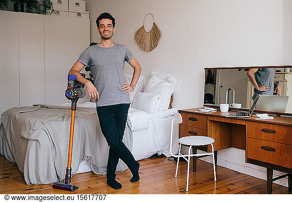 Portrait of confident mid adult man with vacuum cleaner standing in bedroom