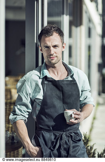 Portrait of confident owner with coffee cup standing at entrance of cafe