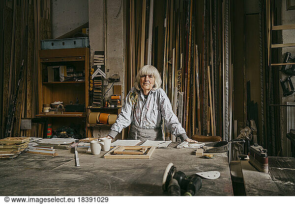 Portrait of confident senior female carpenter leaning on workbench at repair shop