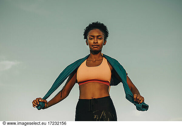 Portrait of confident sportswoman against sky during sunset