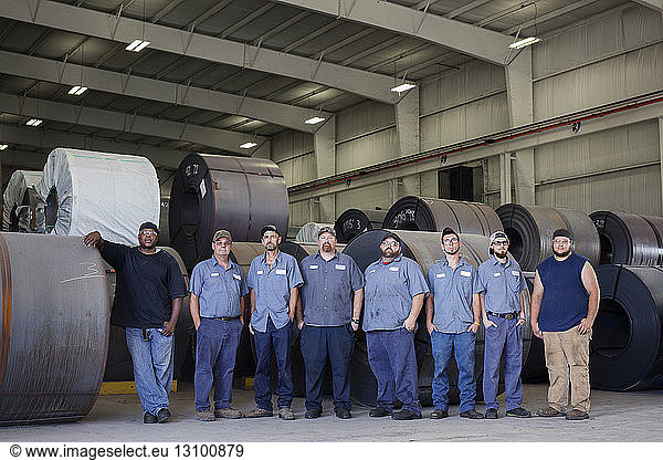 Portrait of confident workers standing against rolled up metal sheets in warehouse