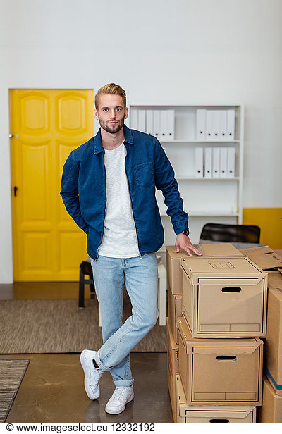 Portrait of confident young businessman standing by cardboard boxes stack at creative office